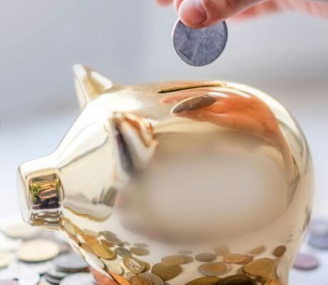 Hand placing coin into a golden piggy bank surrounded by coins, symbolizing savings.