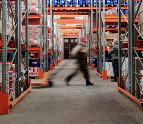 Blurred motion of a man walking in a spacious warehouse aisle between tall shelves.